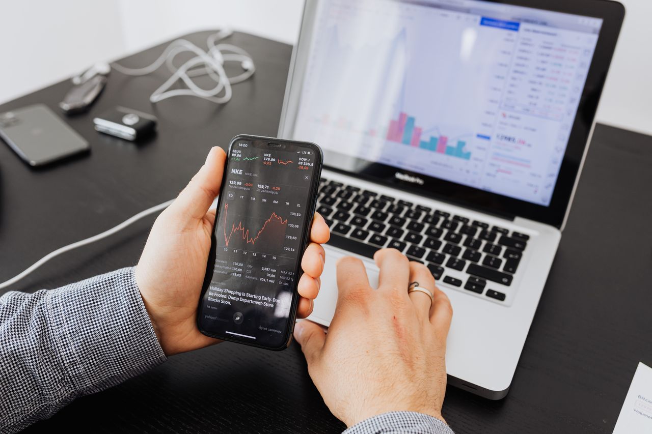 Osoba uvnitř analyzuje akciová data na smartphonu i notebooku.